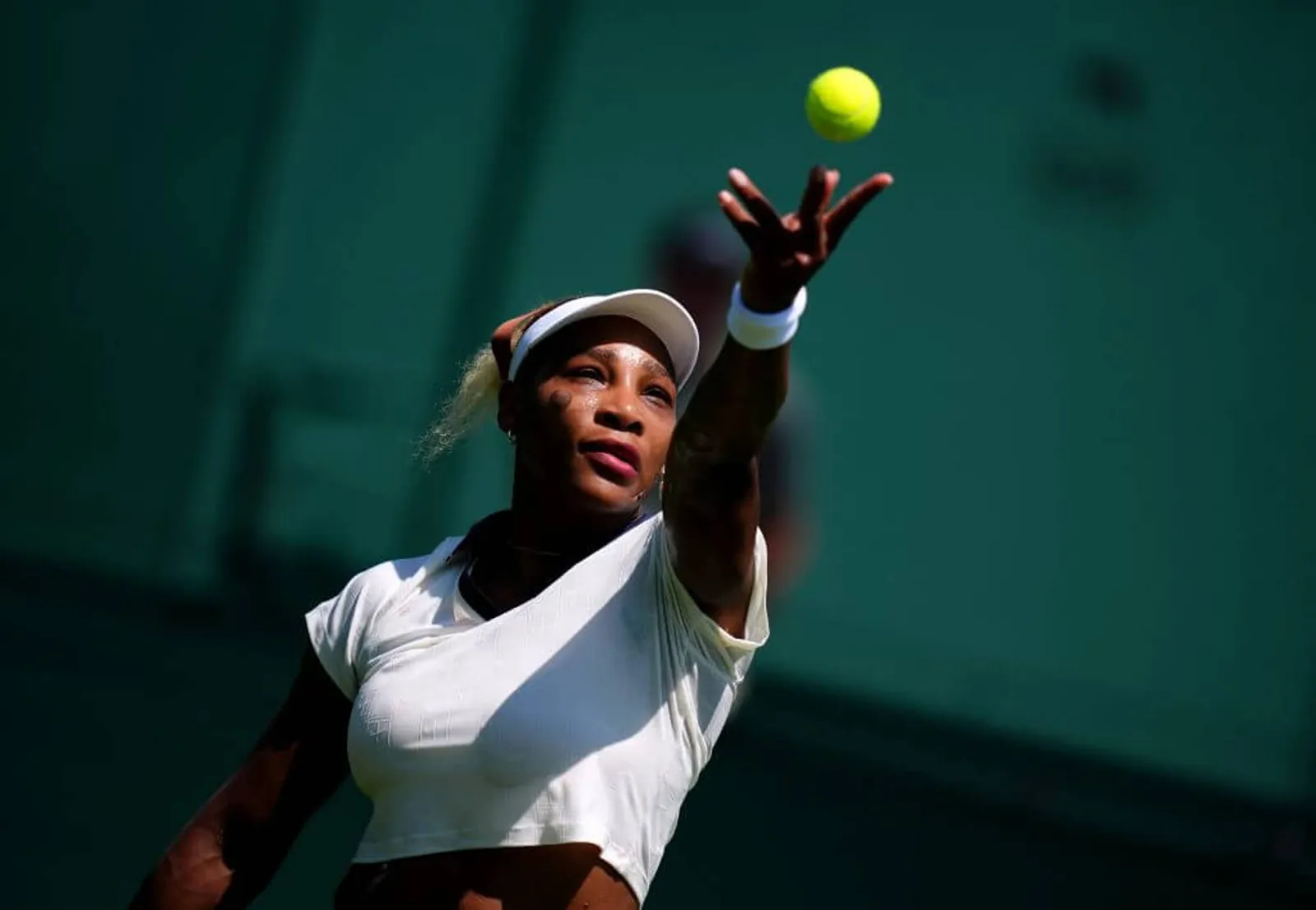 Serena Williams throws a tennis ball into the air on a dark green background wearing all white.
